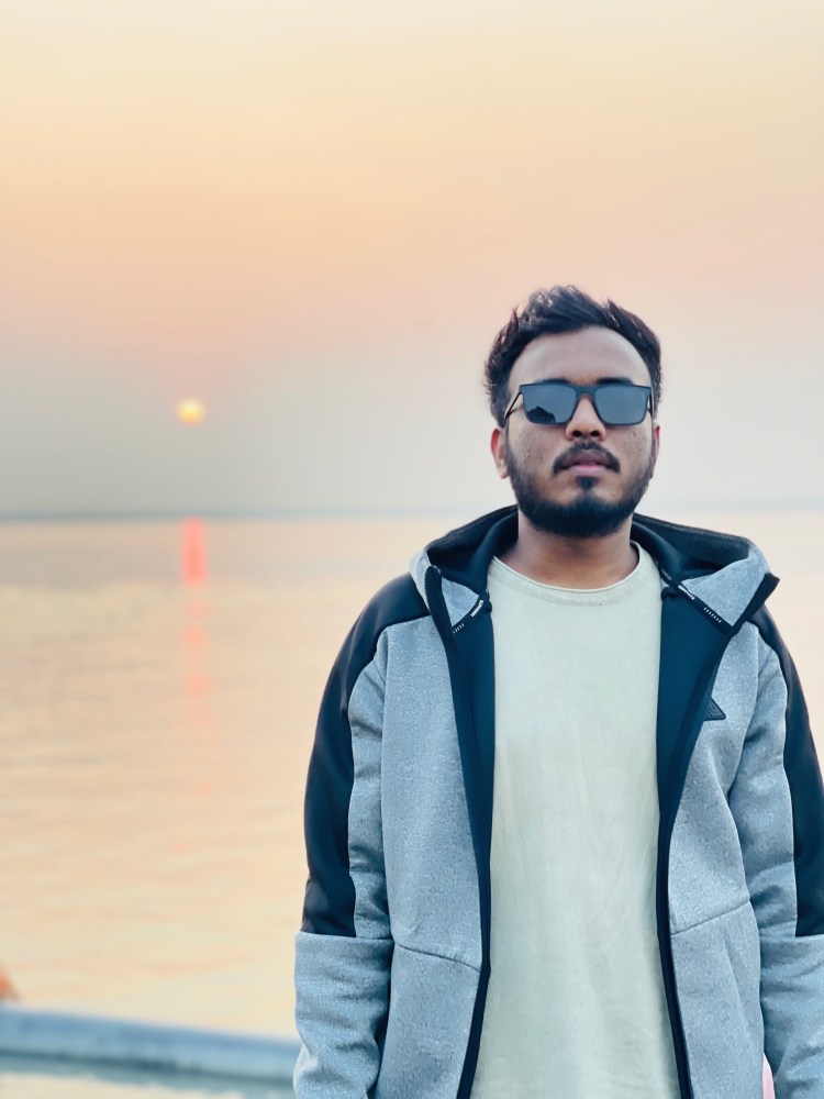 A young man wearing sunglasses and a grey hoodie stands by the water during sunset, with a soft orange sky in the background.